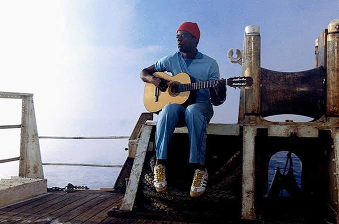 Pele dos Santos in a red cap on the bow of the Belafonte, playing a guitar, open sea behind him