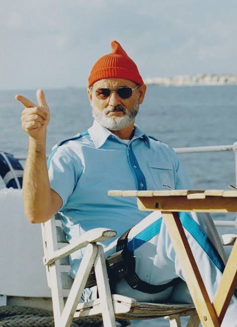 Steve Zissou in a red cap and pale blue shirt, seated on deck, making a point